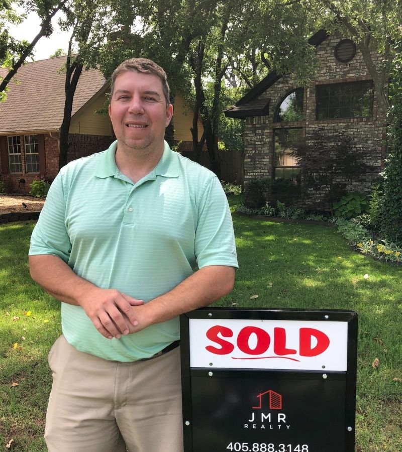 Photo of relocation real estate agent Jimmy Hughes, who is standing next to a "sold" real estate sign in the yard of a house he recently sold. 