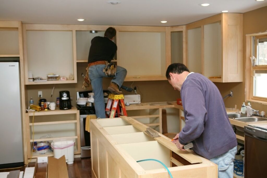 image of two contractors remodeling the inside of a kitchen. They are installing new cabinets and a kitchen island. 